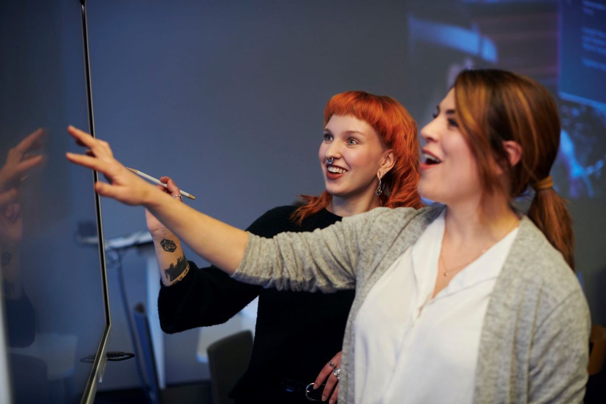 Two young women in a classroom studying something from the screen.
