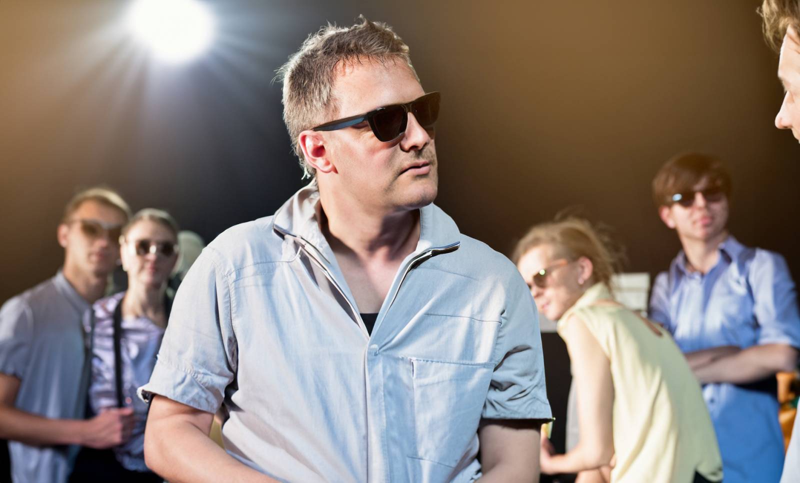 Johtaja, jolla on aurinkolasit silmillään. Tautalla näkyy työryhmän jäseniä. keskustelee henkilön kanssa