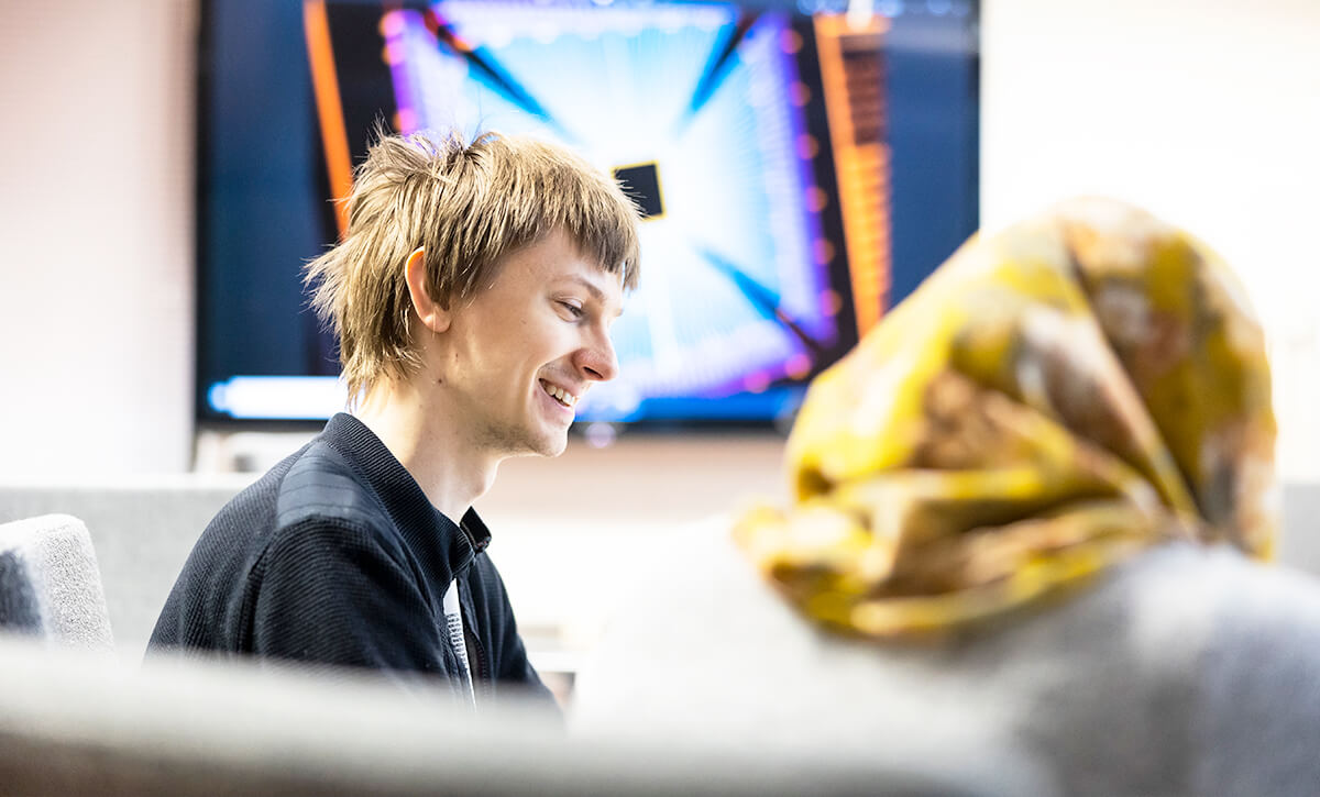 Kaksi opiskelijaa vierekkäin pöydän ääressä. Sivuprofiilista päin näkyvä mies hymyilee. Takaapäin näkyvällä opiskelijalla on päässään huivi.