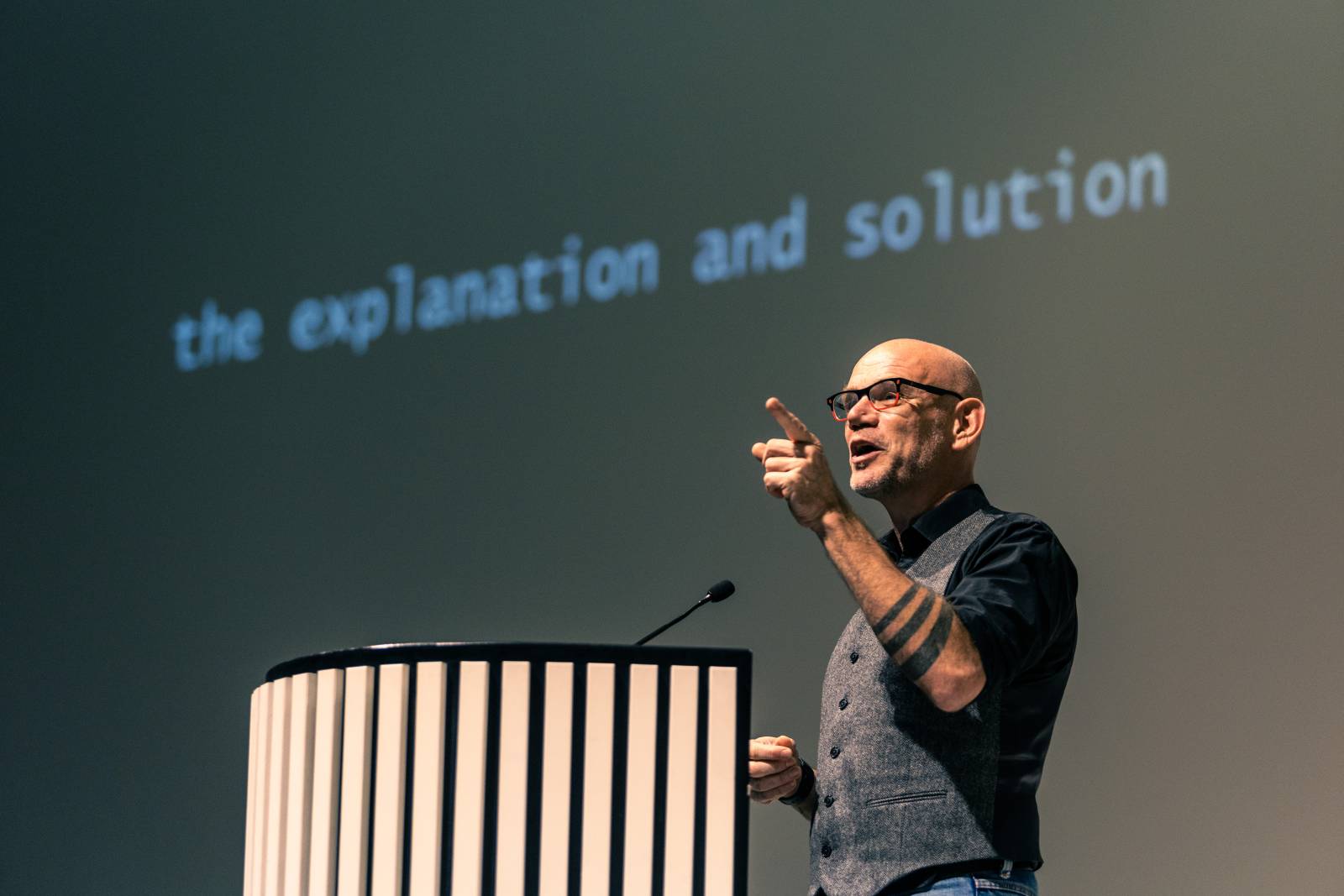 Miesprofessori pitämässä puhetta puhujanpöntössä sormi pystyssä. Taustalla teksti "the explanation and solution"