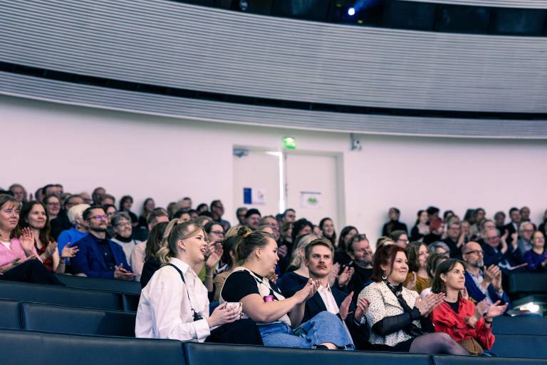 Opiskelijoita ja muita osallistujia istumassa juhlasalissa penkeillä.