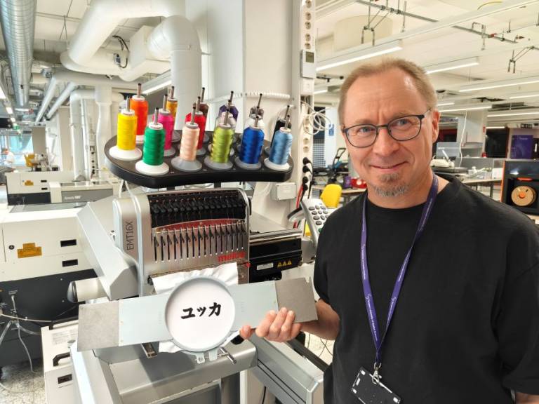Kuvassa hymyilevä Jukka seisoo brodeerauskoneen edessä ja näyttää brodeeraamaansa tekstiä.