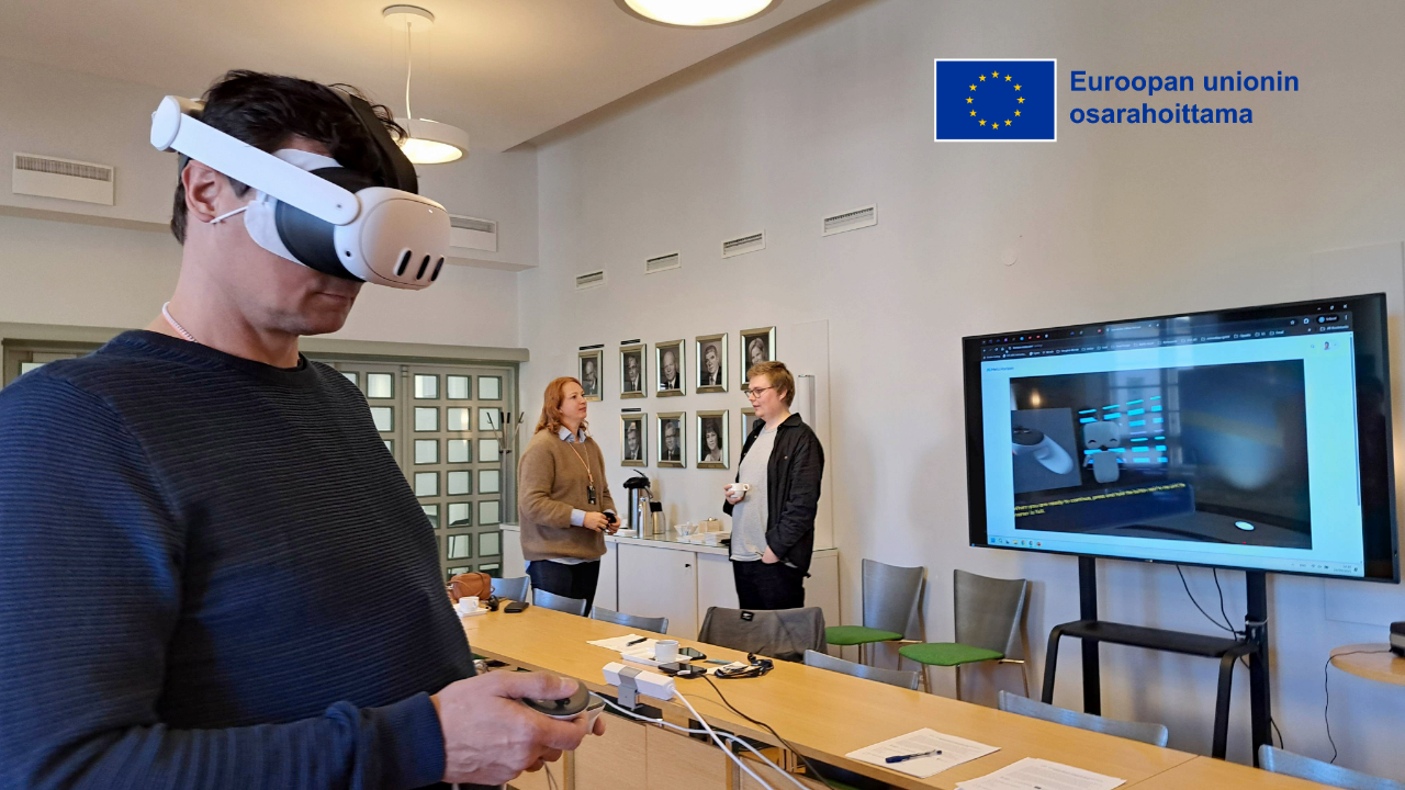 Miehellä on päässä XR-lasit ja käsissä ohjaimet. Taustalla näkyy kaksi muuta henkilöä keskustelemassa tilavassa huoneessa, sekä suuri televisio, mistä näkyy miehen näkemä virtuaalitodellisuus.