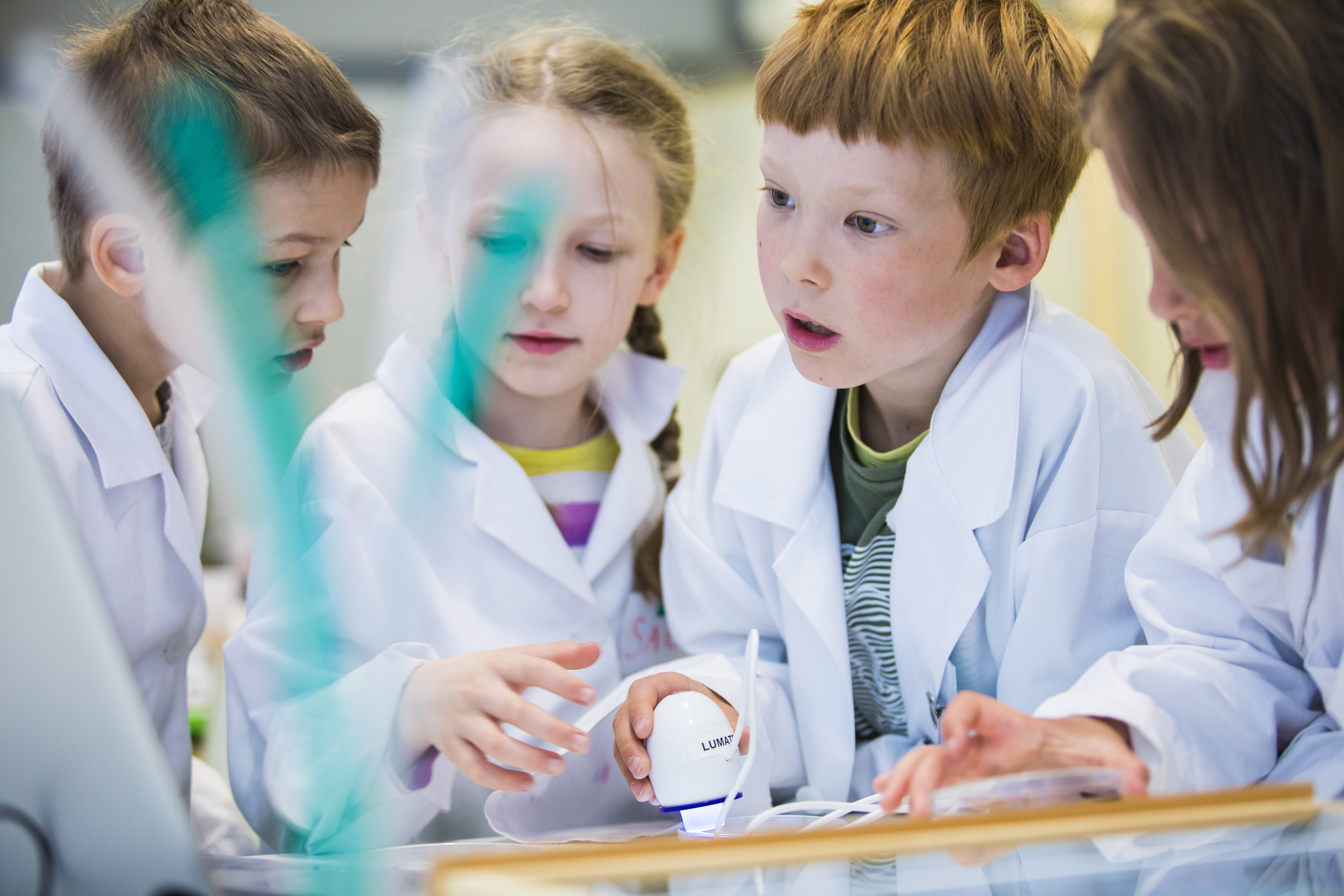 Valokuva, jossa neljä lasta tutkimassa yhdessä näytettä mikroskoopilla laboratoriotakit päällä.