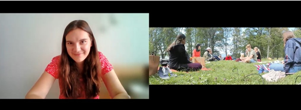 People are sitting on the ground and having picnic, and at the same time one person with long hair and smile on her face is participating remotely and is looking towards the camera.