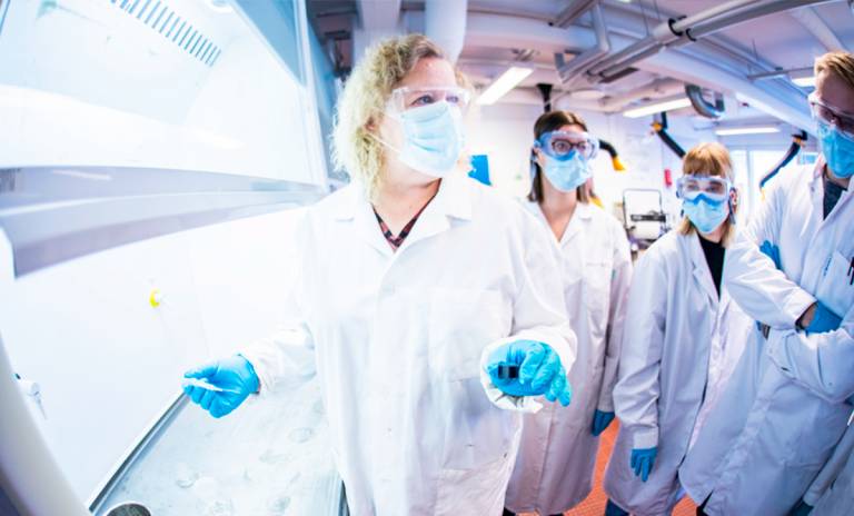 A teacher and three students are wearing masks and safety goggles in a bright laboratory.
