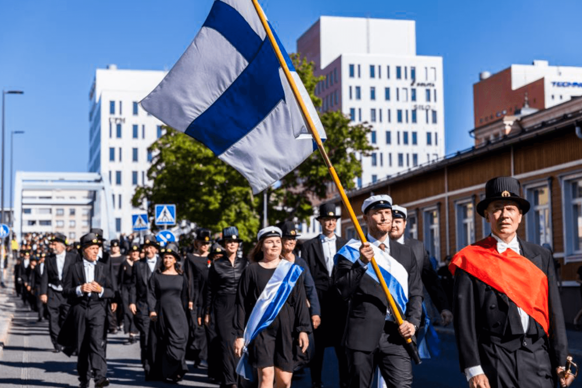 Vuonna 2022 järjestetyn tohtoripromootion kulkue Tampereen kadulla. Kärjessä mies kantaa suomenlippua. Aurinko paistaa.