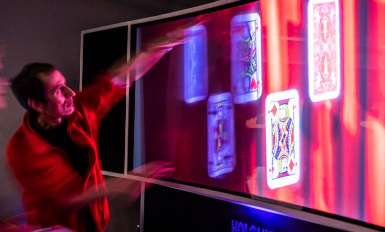 Five big playing cards are displayed on a big screen. A man is standing next to the screen and pointing his hands at the cards.