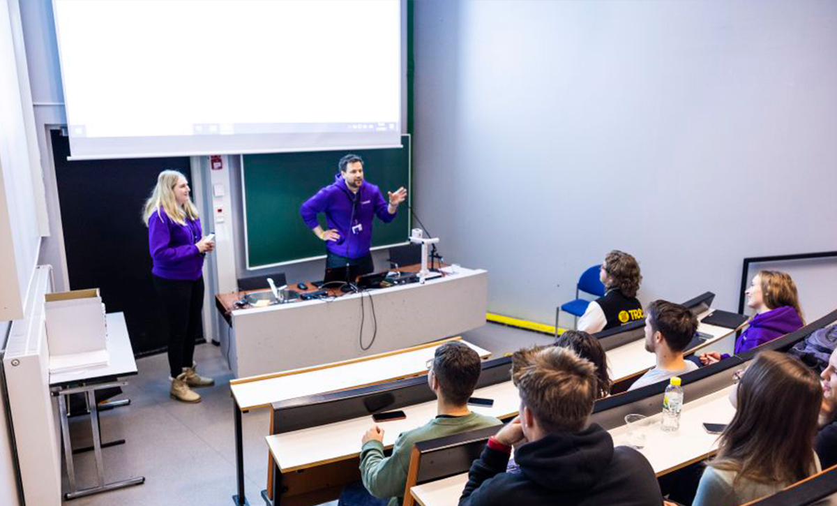 A teaching situation in a small auditorium. The teacher speaks in front with his hand lifted up and the students look at him with interest.