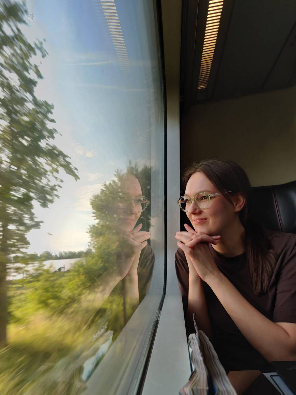 Silmälasipäinen tummahiuksinen nainen katsoo ulos liikuvan junan ikkunasta. 