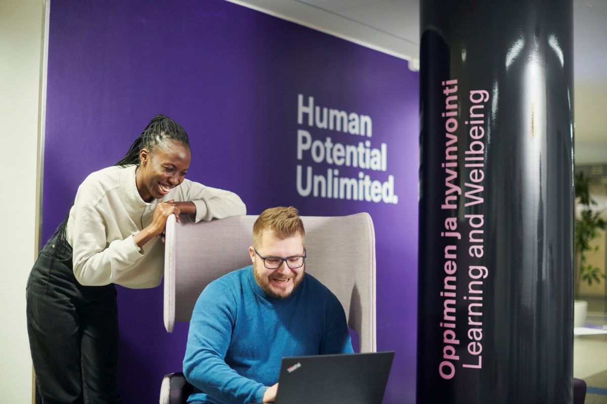 A smiling man sitting on a chair and using his laptop, a girl leaning on the chair in the background.